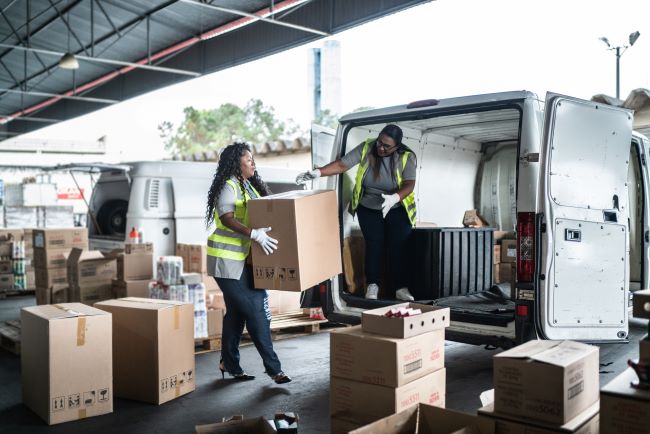 two women in jeans and yellow vests load boxes into a white work van with gloves
